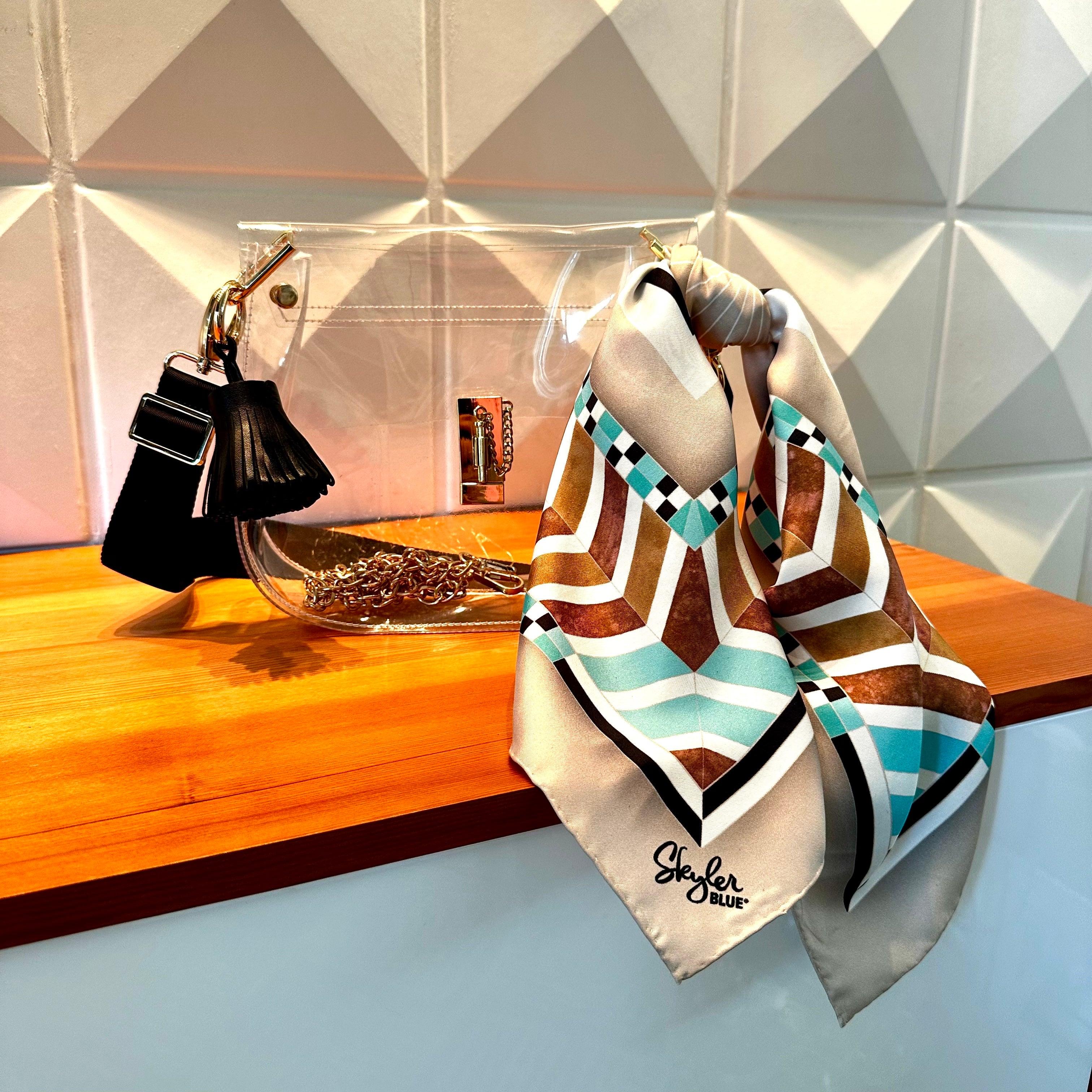 Clear bag with black strap and black tassel with tan, turquoise, brown, and black Aztec print silk scarf tied to purse on wooden surface.