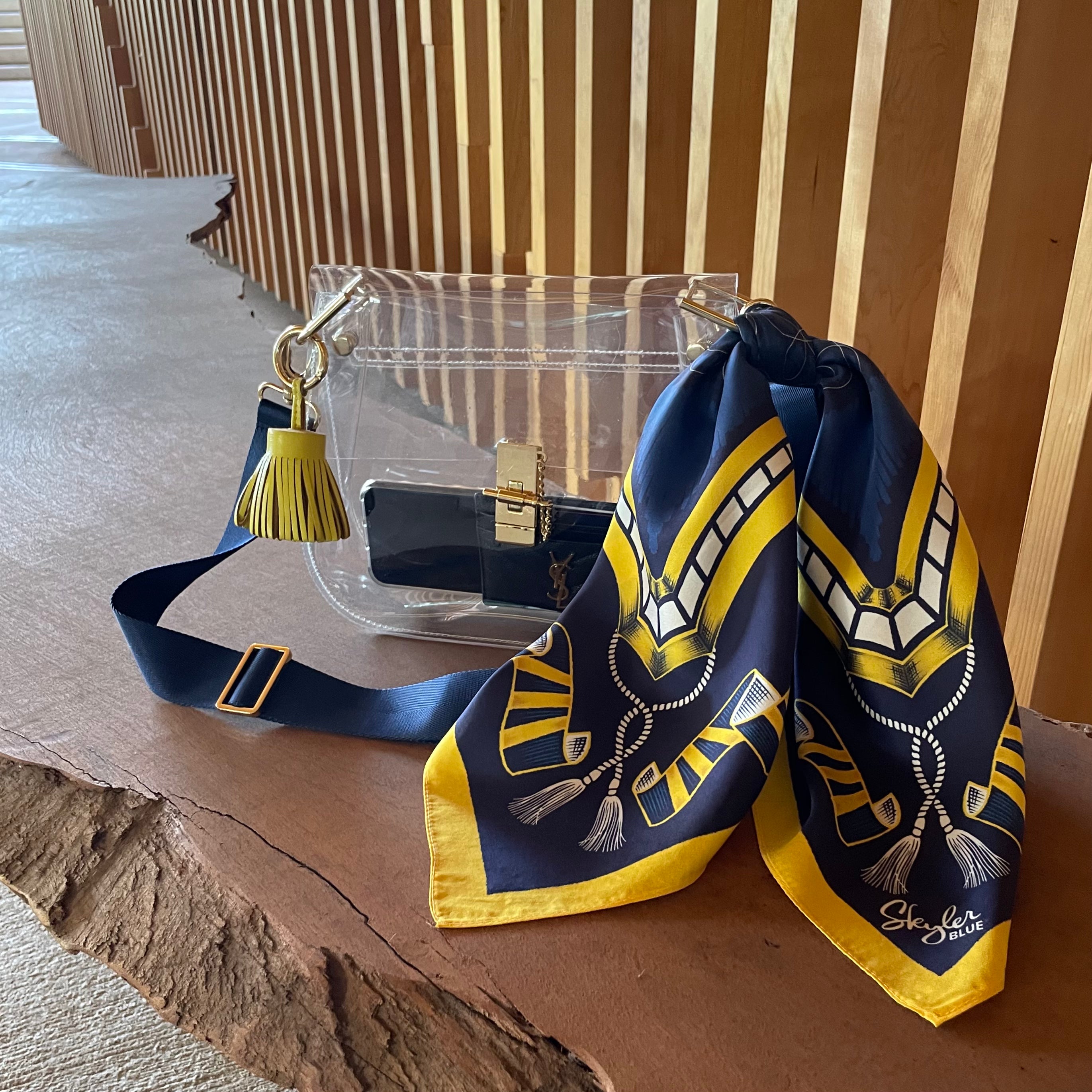 Skyler Blue’s The Ann Arbor Medium Saddle Clear Bag stadium approved clear bag / clear purse including adjustable, nylon webbing shoulder or crossbody strap with herringbone weave and gold hardware, 60-centimeter 100% silk twill scarf, and 100% genuine leather tassel.