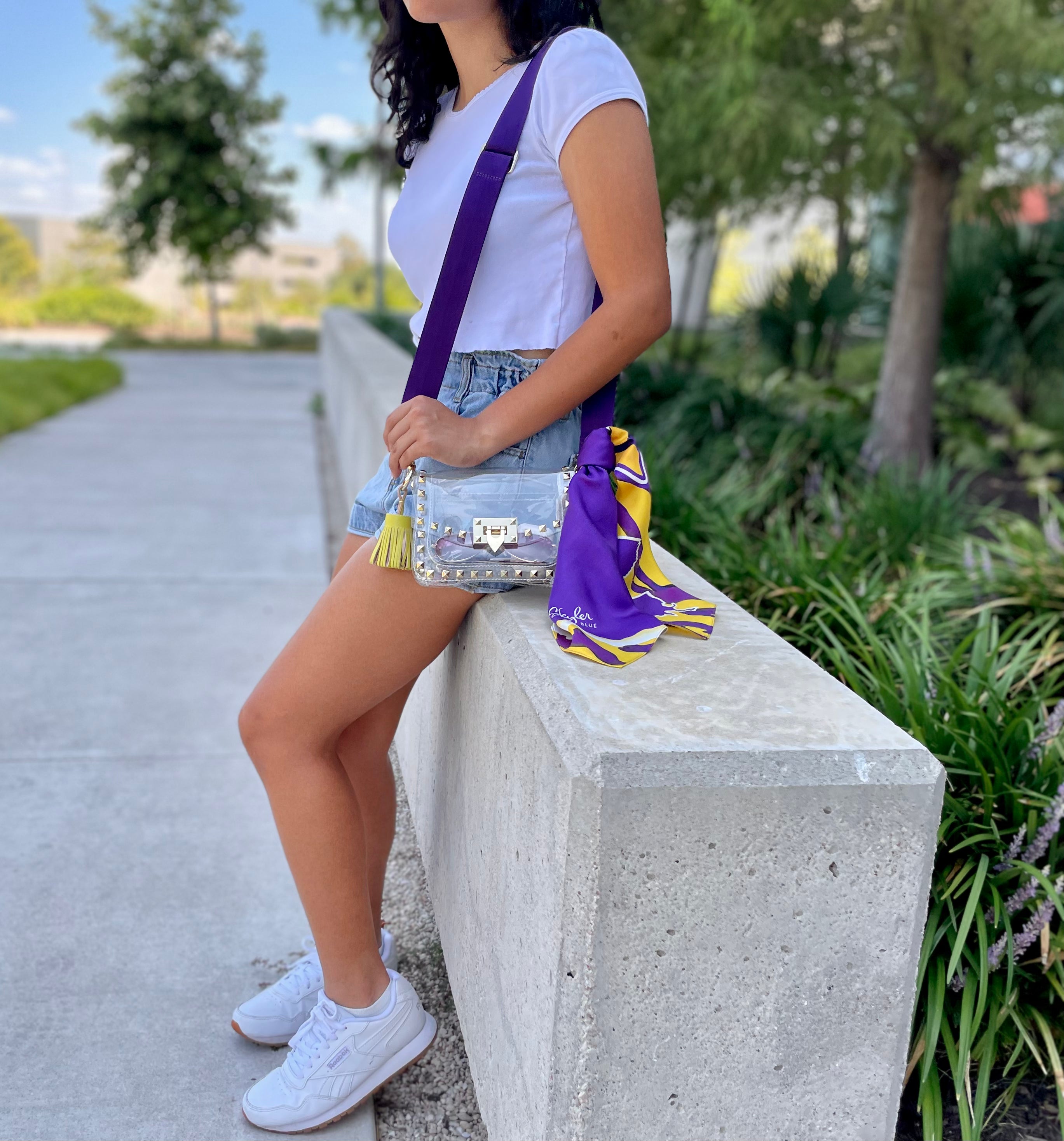 Model wearing Skyler Blue’s The Baton Rouge Small Studded Clear Bag stadium approved clear bag / clear purse including adjustable, nylon webbing shoulder or crossbody strap with herringbone weave and gold hardware, 60-centimeter 100% silk twill scarf, and 100% genuine leather tassel.