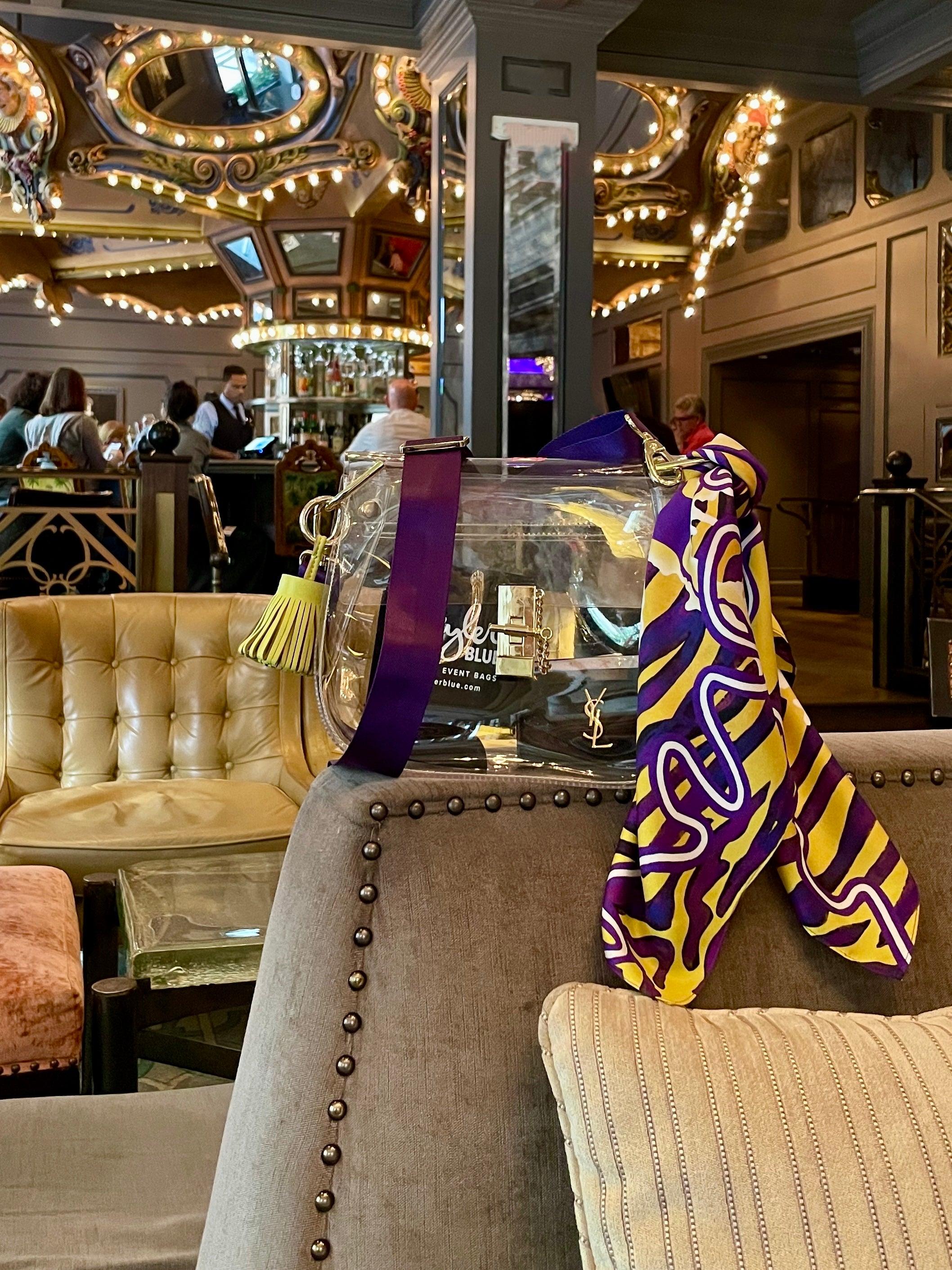 Skyler Blue’s The Baton Rouge Medium Saddle Clear Bag stadium approved clear bag / clear purse including adjustable, nylon webbing shoulder or crossbody strap with herringbone weave and gold hardware, 60-centimeter 100% silk twill scarf, and 100% genuine leather tassel.