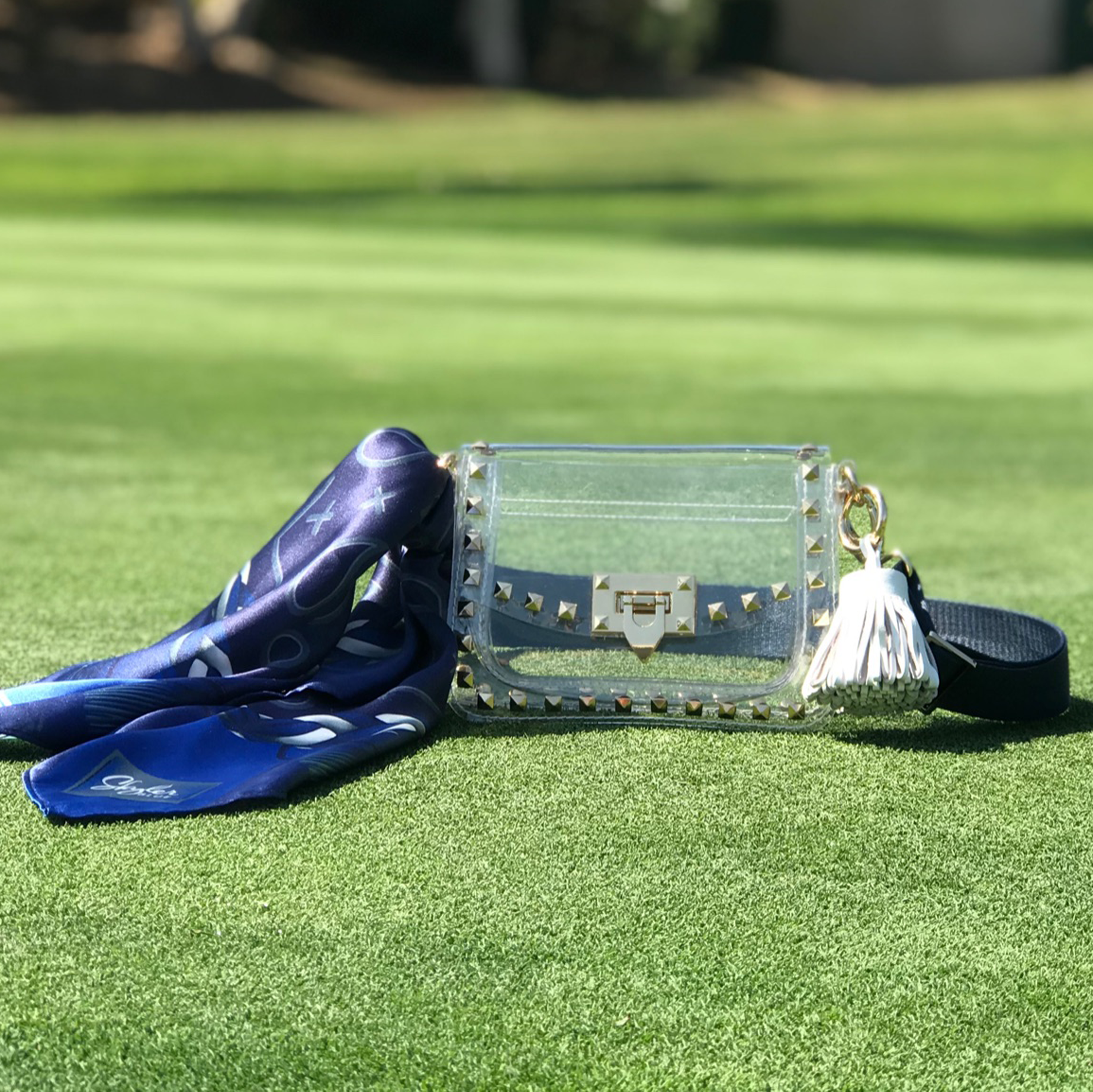 Skyler Blue’s The Dallas 001 Small Studded Clear Bag stadium approved clear bag / clear purse including adjustable, nylon webbing shoulder or crossbody strap with herringbone weave and gold hardware, 60-centimeter 100% silk twill scarf, and 100% genuine leather tassel. 