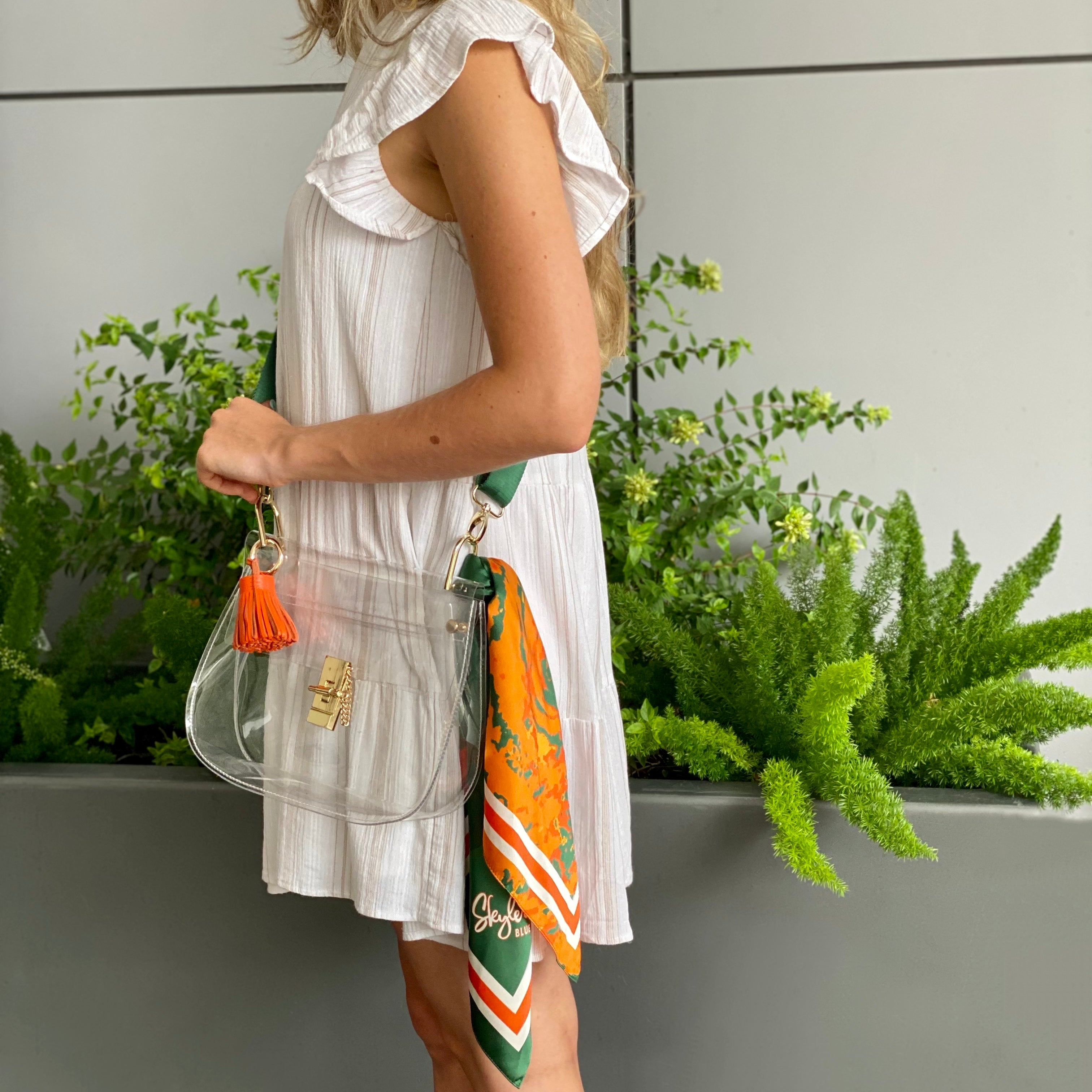Model wearing Skyler Blue’s The Coral Gables Medium Saddle Clear Bag stadium approved clear bag / clear purse including adjustable, nylon webbing shoulder or crossbody strap with herringbone weave and gold hardware, 60-centimeter 100% silk twill scarf, and 100% genuine leather tassel. 