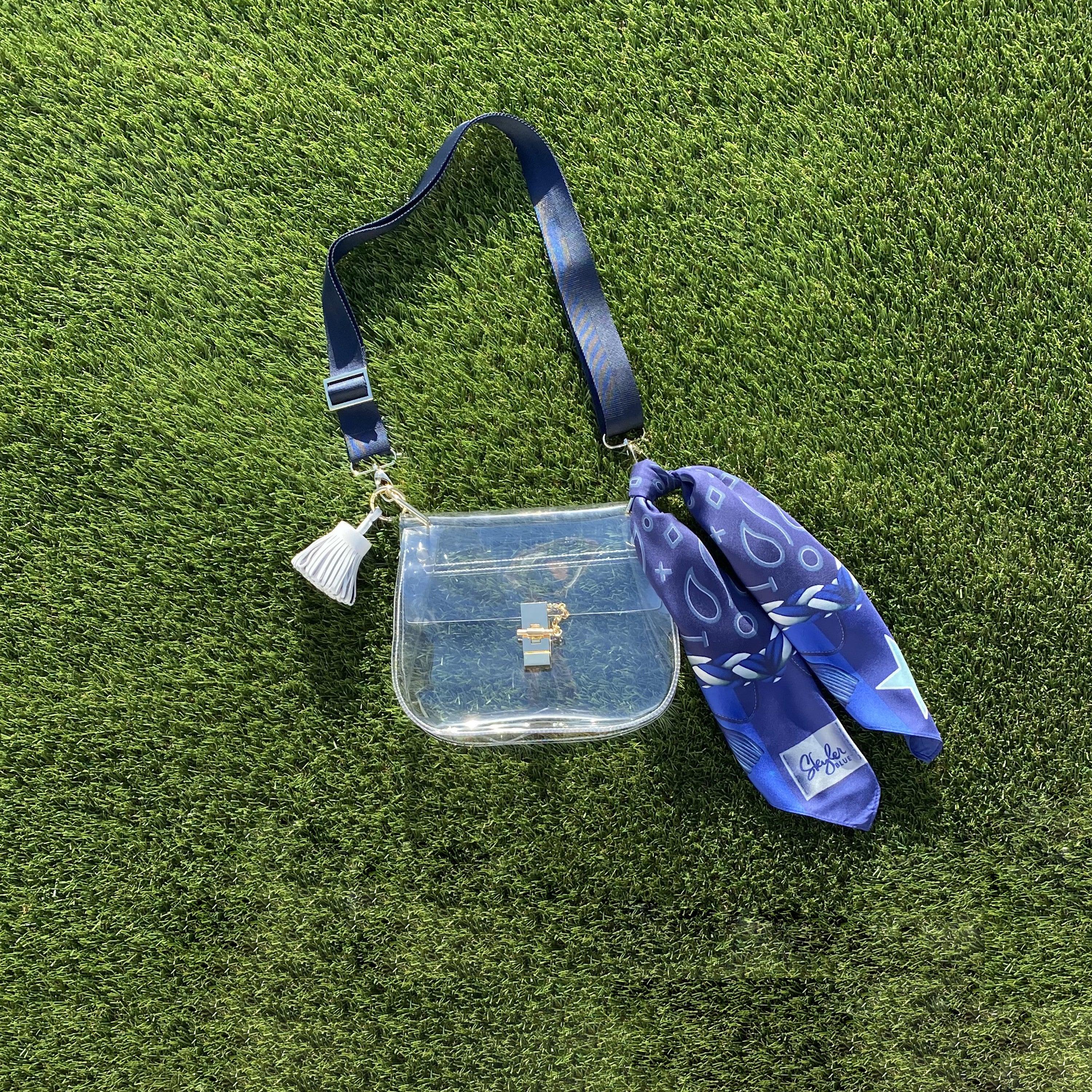 Skyler Blue’s The Dallas 001 Medium Saddle Clear Bag stadium approved clear bag / clear purse including adjustable, nylon webbing shoulder or crossbody strap with herringbone weave and gold hardware, 60-centimeter 100% silk twill scarf, and 100% genuine leather tassel. 