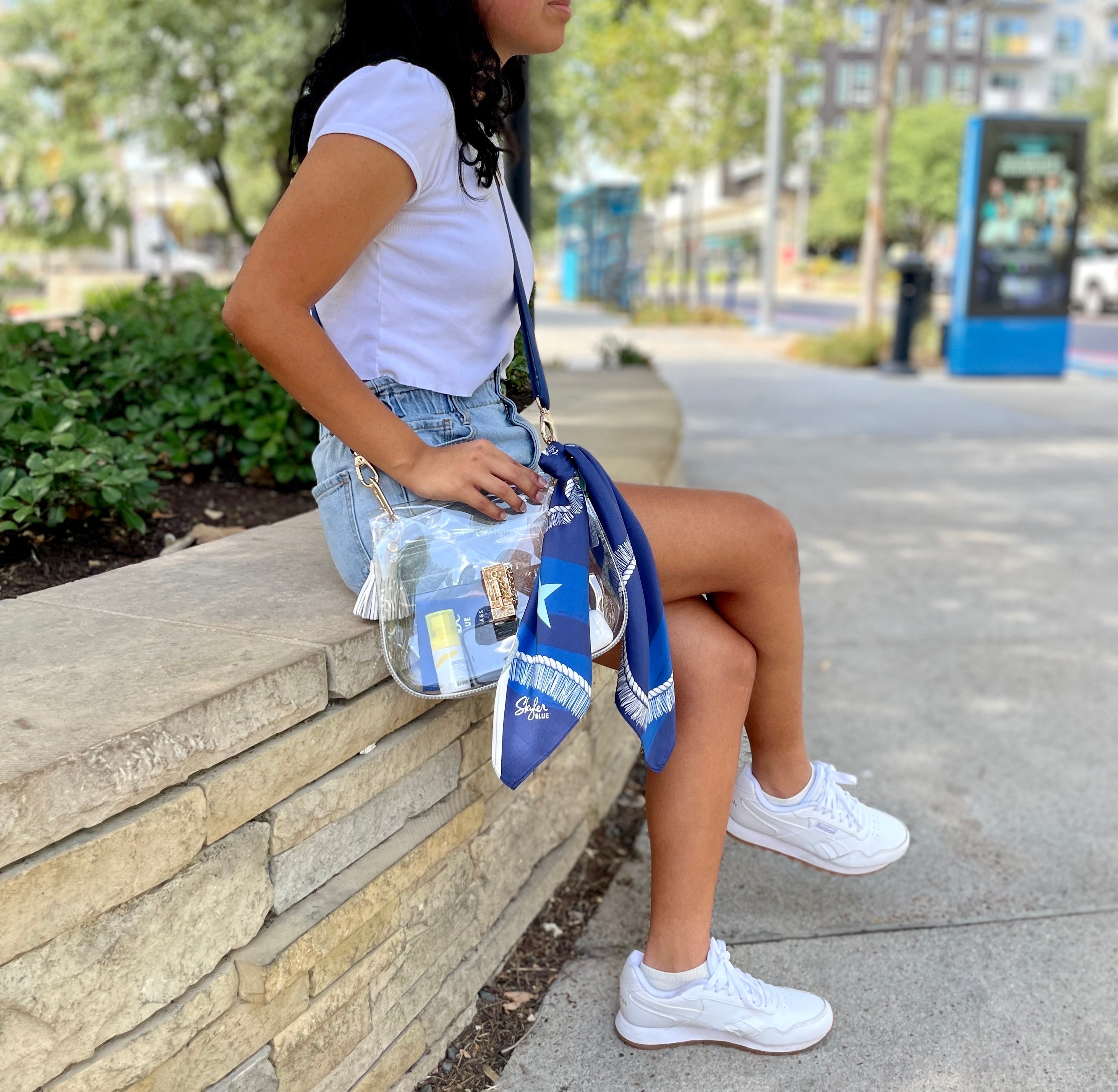Model wearing Skyler Blue’s The Dallas 002 Medium Saddle Clear Bag stadium approved clear bag / clear purse including adjustable, nylon webbing shoulder or crossbody strap with herringbone weave and gold hardware, 60-centimeter 100% silk twill scarf, and 100% genuine leather tassel. 