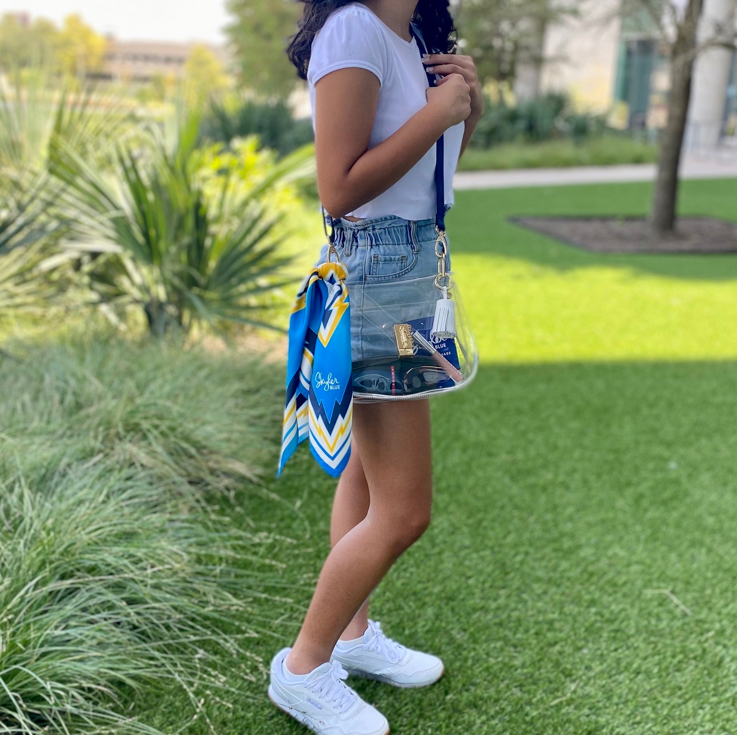 Model wearing Skyler Blue’s The Bolt Medium Saddle Clear Bag stadium approved clear bag / clear purse including adjustable, nylon webbing shoulder or crossbody strap with herringbone weave and gold hardware, 60-centimeter 100% silk twill scarf, and 100% genuine leather tassel. 
