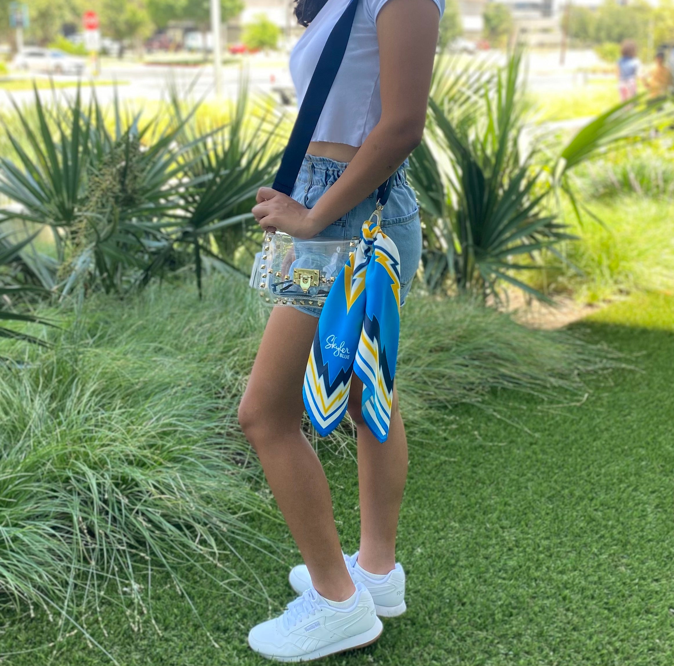 Model wearing Skyler Blue’s The Bolt Small Studded Clear Bag stadium approved clear bag / clear purse including adjustable, nylon webbing shoulder or crossbody strap with herringbone weave and gold hardware, 60-centimeter 100% silk twill scarf, and 100% genuine leather tassel. 