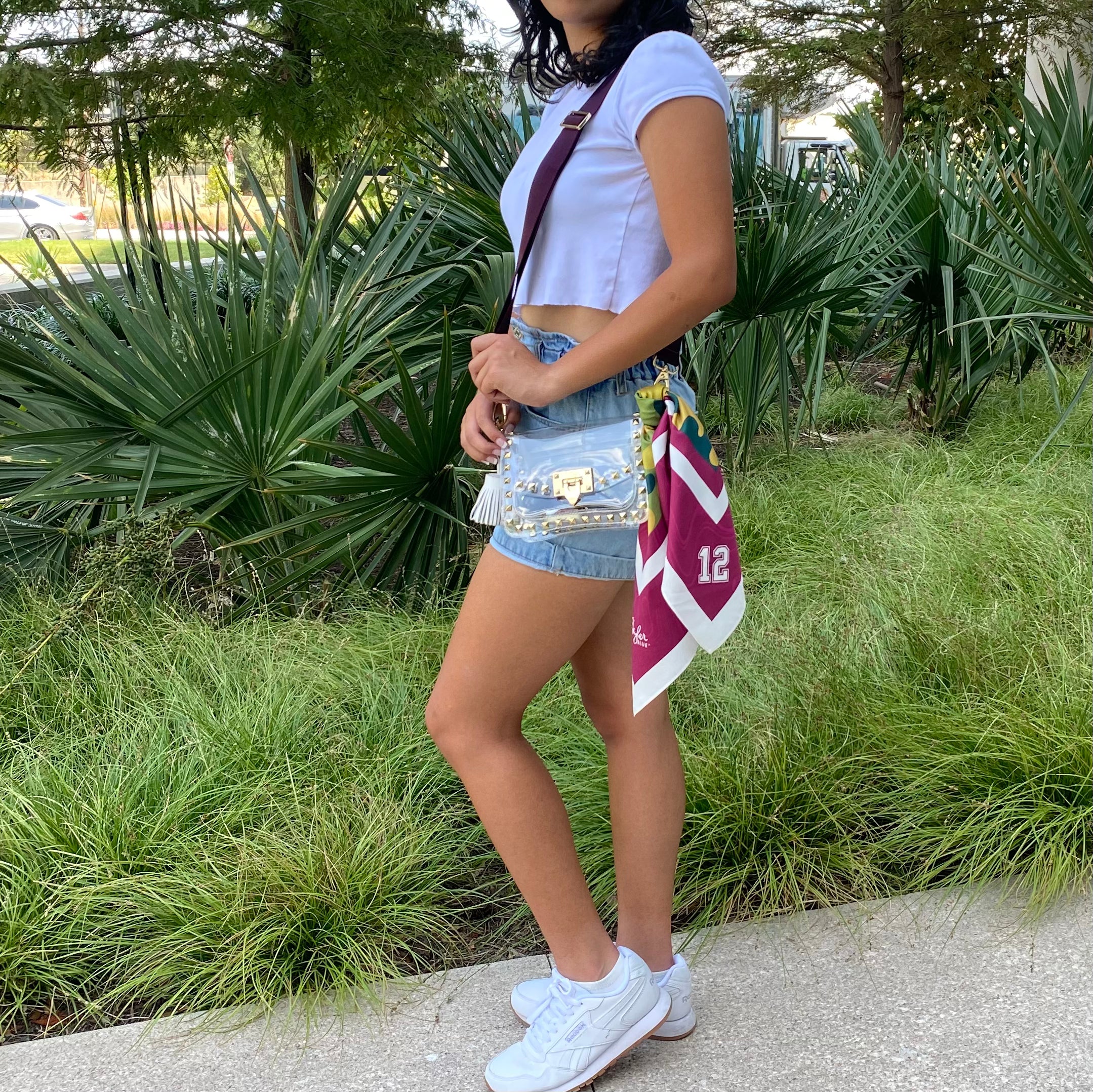 Model wearing Skyler Blue’s The College Station Small Studded Clear Bag stadium approved clear bag / clear purse including adjustable, nylon webbing shoulder or crossbody strap with herringbone weave and gold hardware, 60-centimeter 100% silk twill scarf, and 100% genuine leather tassel. 