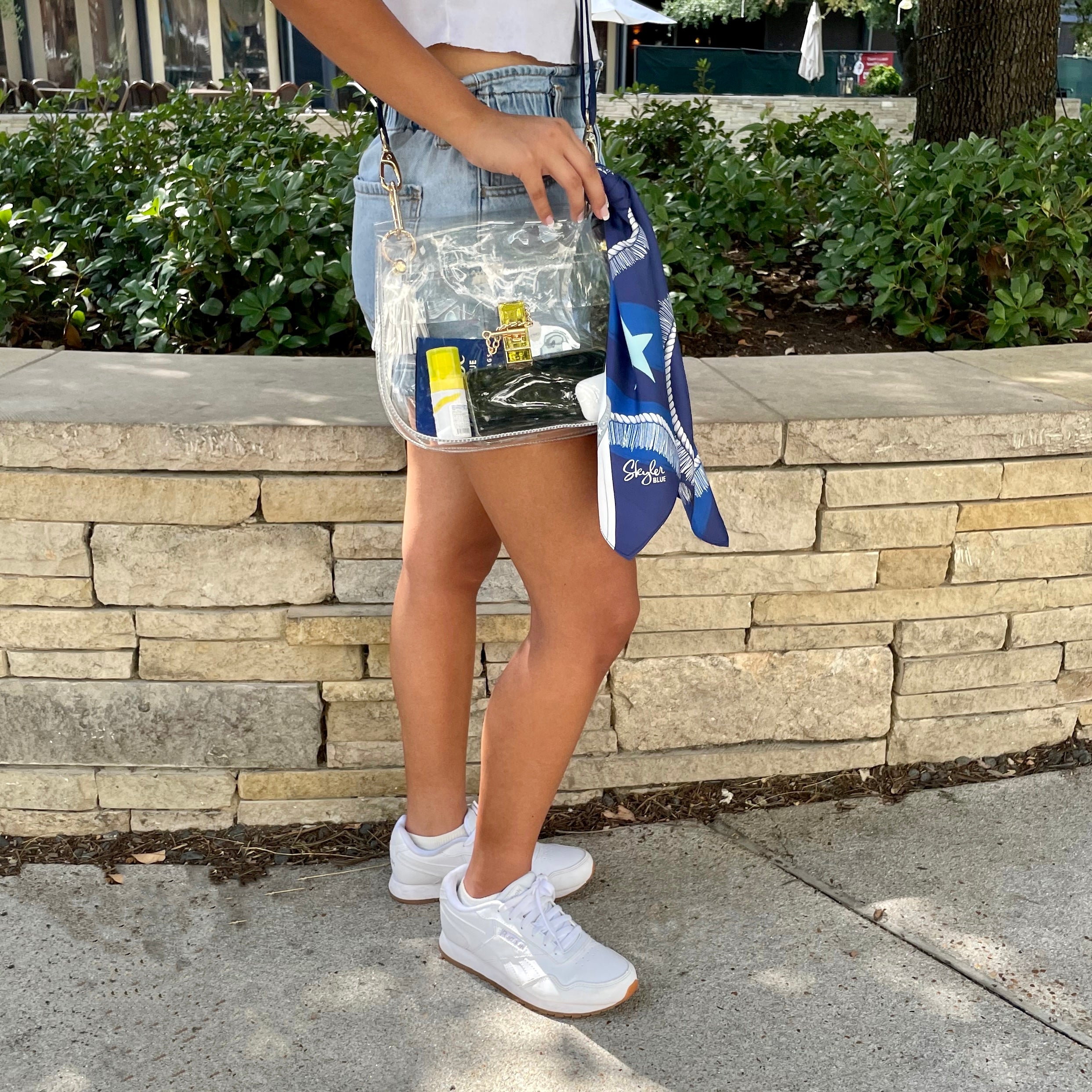 Model wearing Skyler Blue’s The Dallas 002 Medium Saddle Clear Bag stadium approved clear bag / clear purse including adjustable, nylon webbing shoulder or crossbody strap with herringbone weave and gold hardware, 60-centimeter 100% silk twill scarf, and 100% genuine leather tassel. 